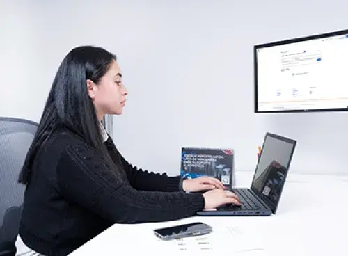 Persona con un bolígrafo en la mano revisando documentos sobre la mesa; a un lado hay un computador portátil.