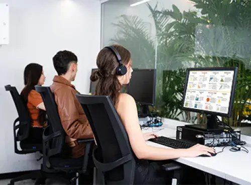 Tres personas sentadas frente a computadoras, una de ellas con auriculares, en una sala con pared de vidrio y plantas al fondo.