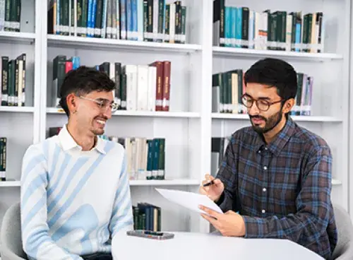 Dos personas observan un documento; una de ellas sostiene un bolígrafo y al fondo hay un estante con libros.
