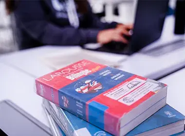 Varios libros sobre la mesa y, al fondo, una persona frente a un portátil.