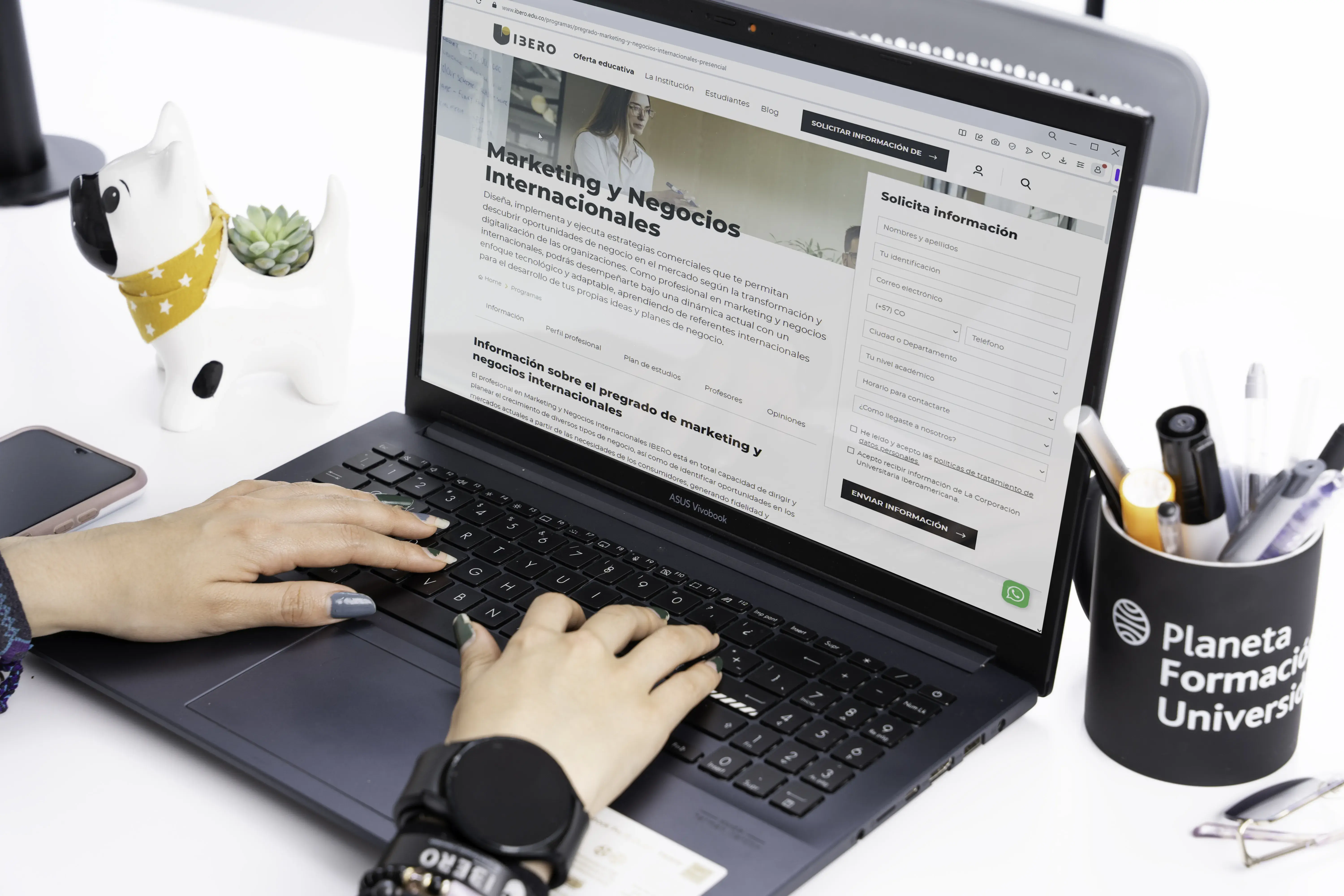 Persona escribiendo en un portátil con la página de un programa universitario de marketing y negocios internacionales abierta; al lado hay un celular, una maceta con forma de perro y un portalápices con bolígrafos.