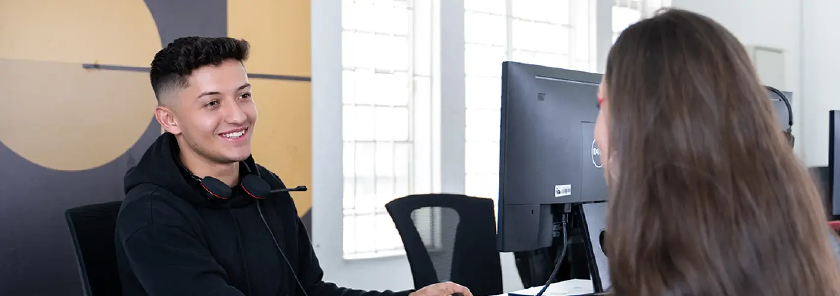 Dos personas hablando,alrededor hay computadores y en la pared hay un cuadro con un círculo amarillo.