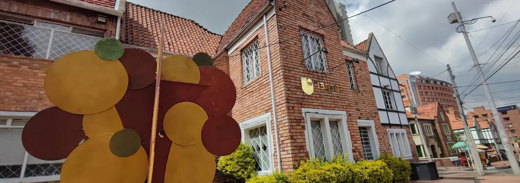 Fotografía de un edificio de ladrillo con techo de teja inclinada y ventanas con rejas blancas de estilo inglés. En primer plano se aprecia una escultura urbana compuesta por discos metálicos en tonos ocres y rojizos, situada en un andén amplio con jardines bien cuidados.